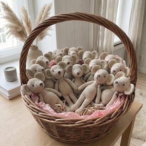 Pode incluir: Uma grande cesta de vime tecida cheia de numerosas bonecas de rato de crochê bege. As bonecas têm orelhas grandes e estão sobre papel de seda rosa. A cesta está sobre uma mesa de madeira, com um vaso de flores secas ao fundo.