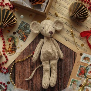 Puede incluir: Una mu&ntilde;eca de rat&oacute;n de ganchillo hecha a mano en hilo beige. El rat&oacute;n tiene orejas grandes, una cola larga y una nariz marr&oacute;n. La mu&ntilde;eca se exhibe sobre una superficie de madera con ef&iacute;meras vintage, incluyendo partituras, sellos y decoraciones doradas.