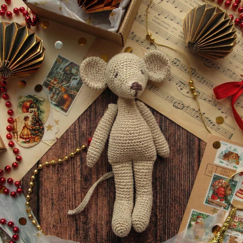 Puede incluir: Una mu&ntilde;eca de rat&oacute;n de ganchillo hecha a mano en hilo beige. El rat&oacute;n tiene orejas grandes, una cola larga y una nariz marr&oacute;n. La mu&ntilde;eca se exhibe sobre una superficie de madera con ef&iacute;meras vintage, incluyendo partituras, sellos y decoraciones doradas.