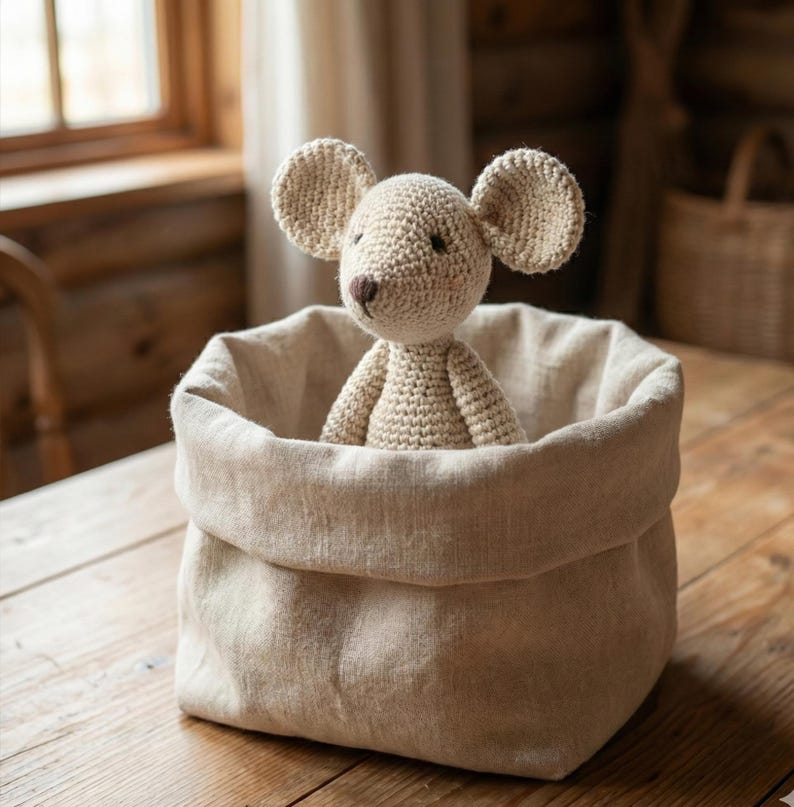 Puede incluir: Un juguete de rat&oacute;n de ganchillo beige con orejas grandes est&aacute; dentro de una cesta de lino beige. La cesta est&aacute; sobre una mesa de madera. El rat&oacute;n tiene una nariz marr&oacute;n y ojos negros peque&ntilde;os. El fondo incluye una ventana y una pared de madera.