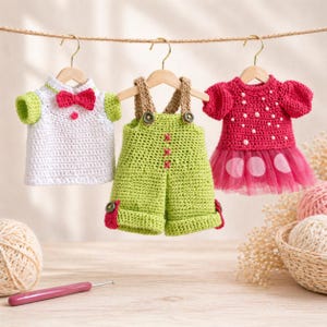 May include: Three handmade crochet baby outfits hanging on a clothesline. The outfits include a white shirt with a red bow tie, green overalls, and a pink dress with a tulle skirt. The outfits are displayed on small wooden hangers.