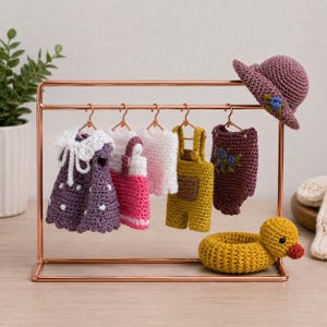 Op de afbeelding: Een kledingrek van roségoudkleurig metaal met vijf gehaakte miniatuurkledingstukken, waaronder een paarse jurk, een roze-witte outfit, een wit topje, een gele tuinbroek en een donkerpaarse romper, met een bijpassende hoed en een gele eend.