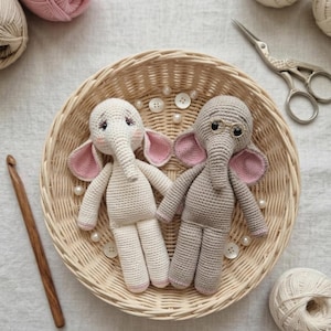 Puede incluir: Dos muñecos de elefante de ganchillo en una cesta de mimbre. Uno es blanco roto, el otro gris, ambos con orejas forradas en rosa. El elefante blanco tiene largas pestañas. El elefante gris lleva gafas redondas pequeñas. La cesta está rodeada de materiales de manualidades.