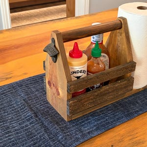 Puede incluir: Un organizador de condimentos de madera teñida oscura con un abridor de botellas de metal integrado en el lateral. El soporte contiene dos botellas exprimibles con tapas roja y verde, una botella de salsa grande y papel de cocina blanco.