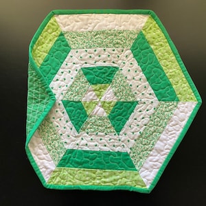 May include: A green and white quilted table runner with a shamrock pattern. The runner is hexagonal in shape and has a geometric design in the center.