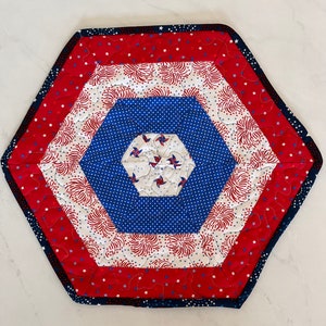 May include: A hexagonal table runner with a red, white, and blue patriotic design. The center features a blue hexagon with white stars and red, white, and blue pinwheels. The outer edges are trimmed with a black and white border.