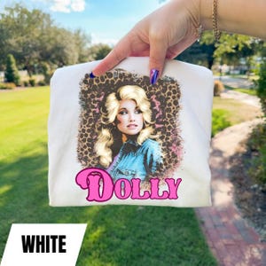 May include: White t-shirt featuring a portrait of a woman with blonde hair and a denim jacket, set against a leopard print background. The word "DOLLY" is printed in pink script. The word "WHITE" is printed in a white rectangle at the bottom.