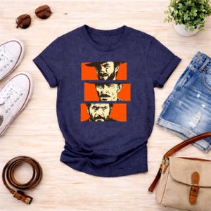 May include: Navy blue t-shirt featuring a graphic of three faces in orange rectangles. The faces appear to be of the same person, with a hat in the top rectangle. The shirt is paired with denim shorts, sunglasses, and a purse.