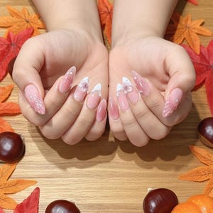 May include: A close-up of a pair of hands with long, almond-shaped, manicured nails. The nails feature a gradient pink color, white tips, and delicate floral and glitter accents. The nails are set against a wooden surface with autumn leaves and chestnuts.