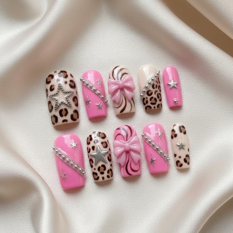 May include: A set of ten artificial nails in various designs. The nails feature pink, leopard print, and cream colors. Some nails have silver star and bow accents. The nails are arranged on a white satin surface.