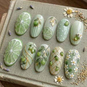 May include: A set of ten almond-shaped, green-themed nail art designs. The designs include lime slices, stripes, polka dots, gold accents, and floral patterns. The nails are arranged on a textured, neutral-toned surface.