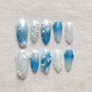 May include: A set of ten press-on nails featuring a gradient of white and blue tones. The designs include hand-painted white line art, shimmering blue glitter accents, and small white spherical beads applied to the surface of several nail tips.
