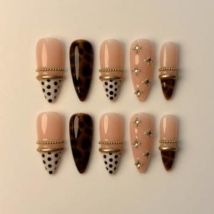 May include: A set of ten artificial nails in various designs. Some nails are nude with gold accents and black polka dots, others feature a tortoiseshell pattern, and some have small star-shaped embellishments.