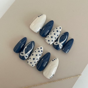 May include: A set of almond-shaped false nails in a navy blue and white color scheme. Designs include polka dots, bow accents, and solid navy blue. The nails are arranged on a neutral surface.