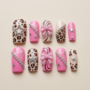 May include: A set of ten artificial nails in various designs. Some nails feature pink polish with silver star accents, while others have a leopard print pattern. Several nails have a bow or star embellishments.