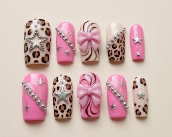 May include: A set of ten artificial nails in various designs. Some nails feature pink polish with silver star accents, while others have a leopard print pattern. Several nails have a bow or star embellishments.