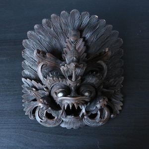 Può includere: Una maschera in legno intagliato a mano di colore marrone scuro, raffigurante una creatura mitica centrale con zanne prominenti, occhi grandi e una cresta a forma di piuma che circonda la testa. L'intaglio in rilievo è dettagliato.