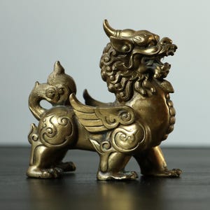 May include: A metallic bronze-coloured figurine of a mythical creature, resembling a winged lion or dragon, featuring intricate swirling patterns on its body, a horned head, an open mouth, and a curled tail, standing in a side profile position.