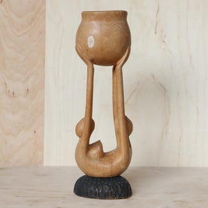 Puede incluir: Escultura de madera tallada que representa una figura sosteniendo un recipiente redondo. La escultura es de madera clara y se apoya sobre una base oscura y texturizada. Los brazos de la figura se extienden hacia arriba, sosteniendo el recipiente.