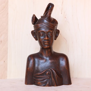 Könnte beinhalten: Dunkelbraune Holzbüste einer Person mit traditionellem Kopfschmuck. Die Skulptur weist detaillierte Gesichtszüge auf und ist mit einem drapierten Gewand über der Brust geschnitzt. Die Büste wird auf einer hellen Oberfläche ausgestellt.