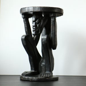 Può includere: Uno sgabello in legno intagliato a mano di colore marrone scuro con una figura centrale in posizione accovacciata. La figura sostiene una piattaforma circolare superiore con le braccia sollevate, mostrando tratti del viso dettagliati.