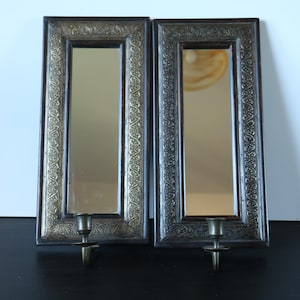 May include: A pair of rectangular wall mirrors with dark wooden frames and ornate metallic borders featuring intricate embossed patterns. Each mirror includes a single attached metal candle holder at the bottom centre, extending outward from the frame.