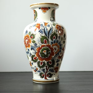 Può includere: Un vaso in ceramica con base color crema decorato con motivi floreali dipinti a mano nei toni dell'arancione, del verde e del blu. Il vaso presenta un bordo svasato, un collo stretto e un corpo arrotondato, rifinito con sottili bande marroni.