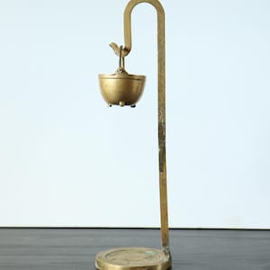 May include: A brass stand with a circular base and a tall, curved arm that supports a small, hanging brass bowl. The bowl features a handle and three small feet, suspended from the hook at the top of the vertical arm, showcasing a metallic finish.