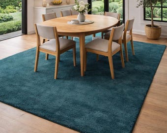 May include: A teal-colored rectangular area rug in a dining room setting. The rug is placed under a wooden oval dining table with six chairs. The room has large windows and a chandelier.