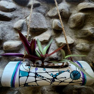 Puede incluir: Una jardinera de bambú blanca pintada a mano con un diseño geométrico en turquesa, morado y negro, suspendida por una cuerda. La jardinera contiene varias plantas verdes y moradas, creando un elemento decorativo natural.