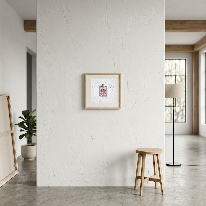May include: A square framed wall art piece featuring a central rectangular illustration of a red and blue patterned garment, set against a white background with scattered blue and grey speckles, enclosed in a light-toned wooden frame.