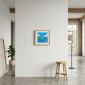 May include: A square framed art print featuring a stylized illustration of a coastal scene with a bright blue sky, a deep blue ocean, and a green shoreline. The artwork is displayed in a light wood frame centered on a white textured wall.