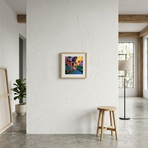 May include: A square framed art print featuring a vibrant, colorful alleyway scene with deep blues, bright yellows, and rich purples. The artwork is centered on a textured white wall, positioned above a wooden stool in a bright, open room.