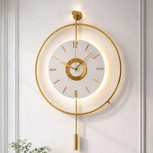 Op de afbeelding: Een moderne wandklok met een goudkleurig cirkelvormig frame en een witte wijzerplaat. De klok heeft gouden wijzers en uurmarkeringen, met een slinger eronder. De klok is bevestigd aan een witte muur. De diameter is ongeveer 30 cm.