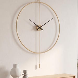 Pode incluir: Um relógio de parede minimalista extra grande, dourado, com 90 cm de diâmetro, ponteiros pretos e um pêndulo. O relógio está montado numa parede branca acima de uma prateleira de madeira com vasos decorativos. O texto "Extra Large" e as dimensões são visíveis.
