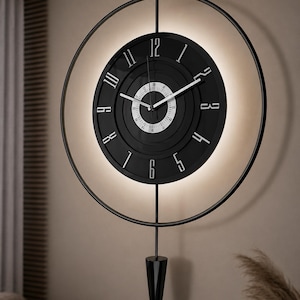 Op de afbeelding: Een moderne zwarte wandklok met een cirkelvormig ontwerp. De wijzerplaat is zwart met witte cijfers en wijzers. Het is omgeven door een zwarte metalen ring en heeft een decoratieve slinger. De klok is van achteren verlicht.