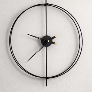 Puede incluir: Un reloj de pared moderno y minimalista con marco y agujas de metal negro. La esfera del reloj es abierta, con dos círculos concéntricos que forman el marco. Las agujas y el círculo central son negros, con detalles dorados.