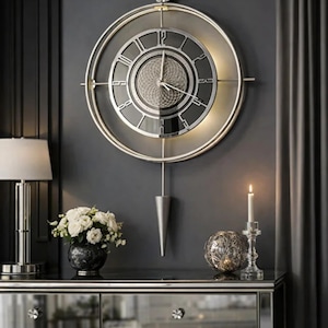 Op de afbeelding: Een ronde wandklok met een zilverkleurige rand, een donkere wijzerplaat met zilveren Romeinse cijfers en een geometrisch patroon in het midden. Een lange, zilverkleurige slinger hangt aan de onderkant en de klok is van achteren verlicht.