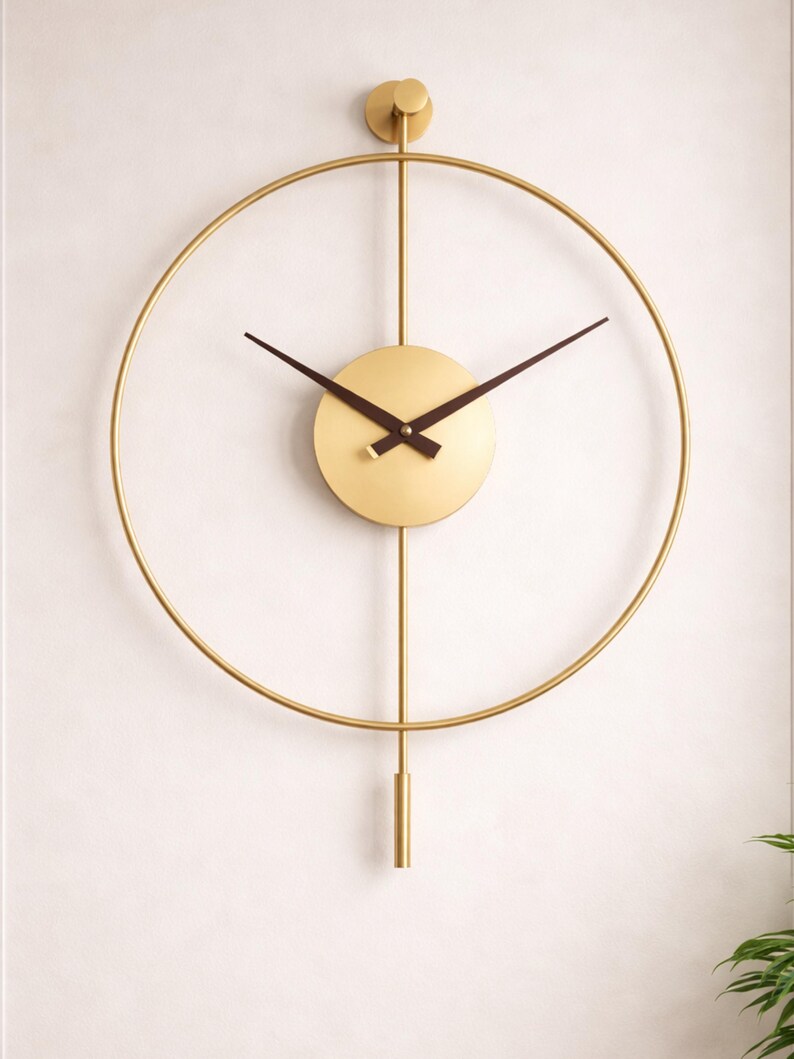 K&ouml;nnte beinhalten: Eine goldfarbene Wanduhr mit minimalistischem Design. Die Uhr hat einen kreisf&ouml;rmigen Rahmen, ein rundes Zifferblatt und schwarze Zeiger. Die Uhr hat ein Gewicht im Pendelstil am unteren Ende und einen kleinen kreisf&ouml;rmigen Akzent oben.