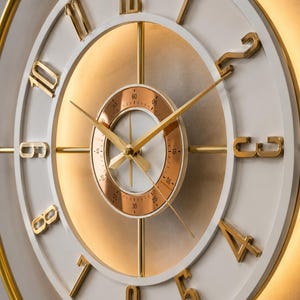 May include: A round wall clock featuring a white face with raised gold-tone numerals from one to twelve. The center includes a smaller copper-toned dial with minute markers and gold hands. A gold outer ring frames the clock with integrated lighting.