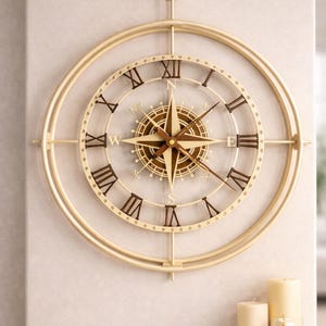 Puede incluir: Un reloj de pared dorado con diseño de brújula. El reloj presenta números romanos y una rosa de los vientos en el centro. El reloj es redondo y tiene un anillo exterior decorativo. El reloj está montado en una pared de color claro.