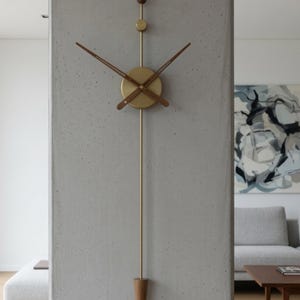 Puede incluir: Un reloj de pared moderno, de 85 cm de altura, con esfera y manecillas de latón. El reloj presenta detalles de madera en las manecillas y un péndulo cónico de madera. El diseño es minimalista y elegante.
