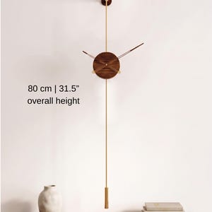 Puede incluir: Reloj de pared moderno con esfera de madera oscura y detalles dorados. El reloj, con péndulo, mide 80 cm de altura total. Está montado en una pared blanca.