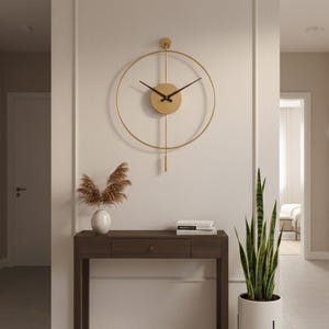 K&ouml;nnte beinhalten: Eine goldfarbene, kreisf&ouml;rmige Wanduhr mit schwarzen Zeigern h&auml;ngt &uuml;ber einem dunklen Holzkonsolentisch. Eine wei&szlig;e Vase mit getrockneten Pflanzen und ein Stapel B&uuml;cher stehen auf dem Tisch. Rechts steht eine hohe Topf-Schlangenpflanze.
