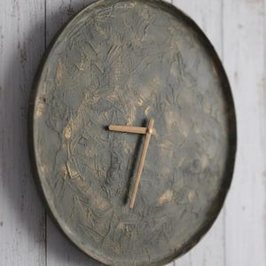 May include: A circular wall clock with a textured, muted green and gold face. The clock's surface has a rough, layered texture. The hands are made of light-coloured wood. The clock is mounted on a white wooden wall.