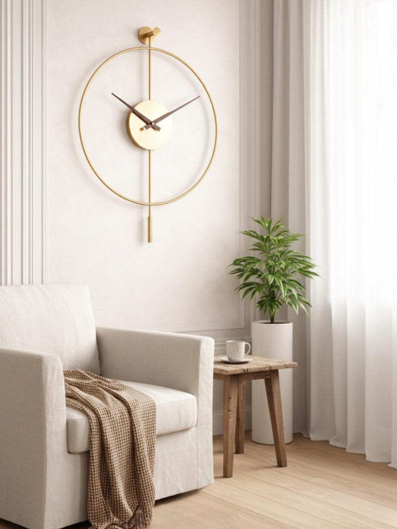 K&ouml;nnte beinhalten: Eine goldfarbene, minimalistische Wanduhr mit kreisf&ouml;rmigem Rahmen und Pendel. Das Zifferblatt ist hellbeige mit schwarzen Zeigern. Die Uhr ist an einer wei&szlig;en Wand befestigt. Der Raum verf&uuml;gt au&szlig;erdem &uuml;ber einen wei&szlig;en Sessel, einen Beistelltisch aus Holz und eine Topfpflanze.