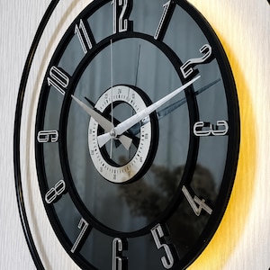 May include: A round wall clock with a black frame and a dark reflective face. It features raised silver-toned numerals from 1 to 12 and a central white circular dial with 24-hour markings. The clock has silver hands and a warm light glow.