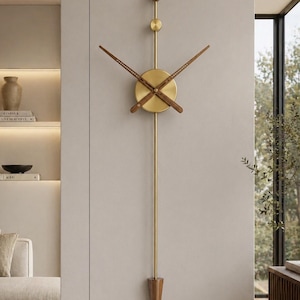 Puede incluir: Un reloj de pared moderno con acabado de latón y detalles de madera. El reloj mide 85 cm (33,5 pulgadas) de largo y presenta una esfera redonda con manecillas de madera. Un péndulo de madera cuelga debajo. El reloj está montado en una pared neutra.