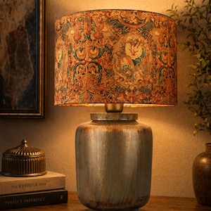 May include: A decorative table lamp with a patterned lampshade in shades of orange, green, and brown. The base is a distressed, metallic blue-gray. The lamp sits on a wooden surface, next to a small decorative box and books.