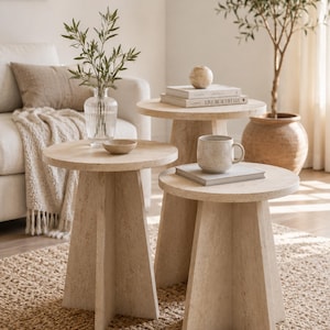 Puede incluir: Tres mesas auxiliares beige con tapas redondas y bases geométricas dispuestas sobre una alfombra tejida. Las mesas varían en altura y están hechas de un material claro y texturizado. Una mesa tiene una taza y un libro, otra un cuenco pequeño y la tercera un jarrón con vegetación.